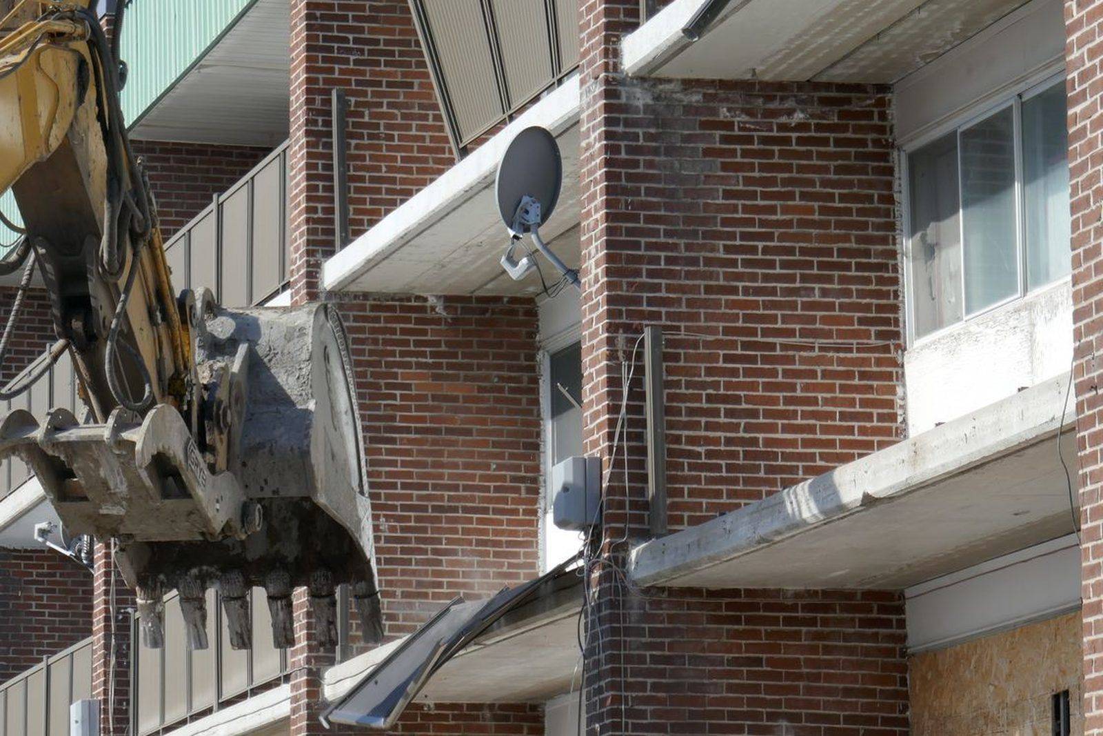 Excavator with a hydraulic thumb attachment pulling apart the balconies of a multi-story brick apartment building during a structural demolition