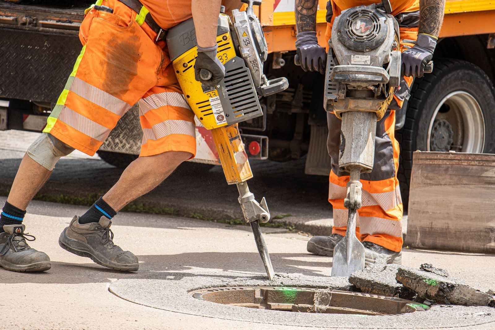 Two workers in high-visibility gear breaking out concrete and asphalt with pneumatic jackhammers around a utility opening in a paved surface