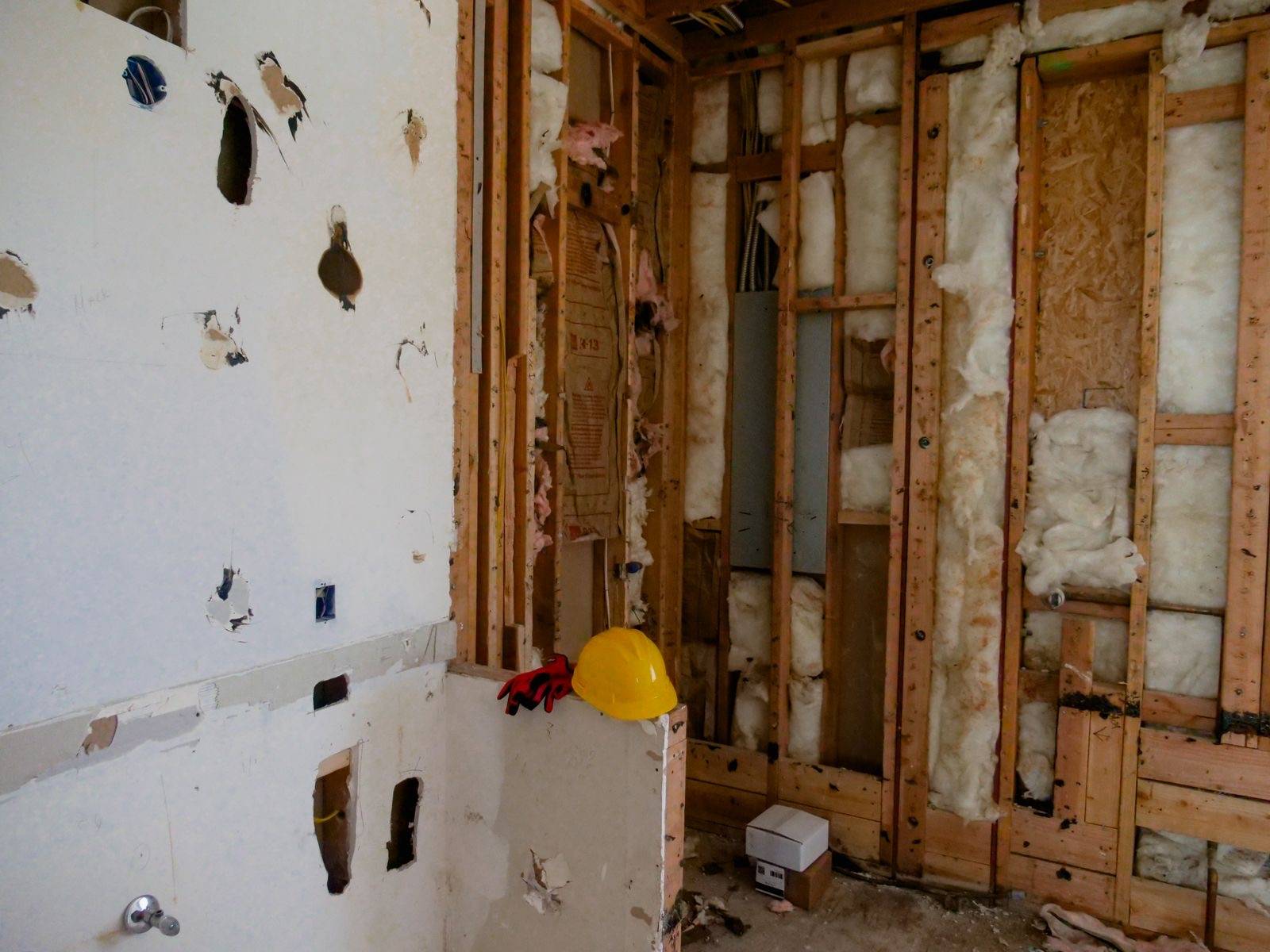 Room stripped to the studs during an interior demolition, with drywall removed, insulation exposed, and a hard hat and work gloves resting on a half wall