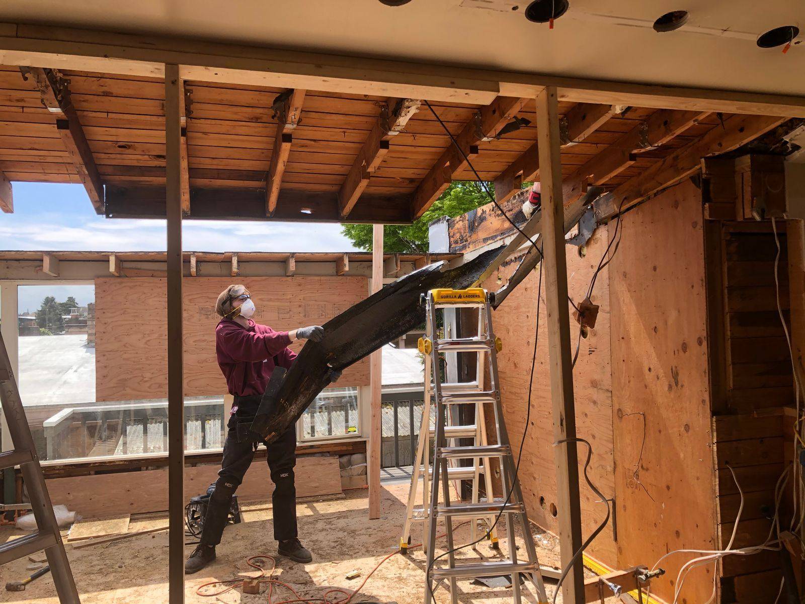 Worker in a respirator pulling down old framing inside a gutted room with the rafters and sheathing exposed during a selective demolition