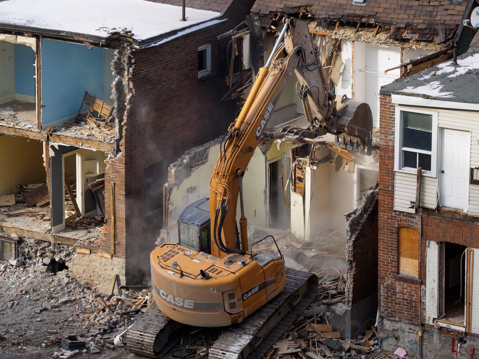 Tracked excavator working inside the gutted shell of a brick house during a residential demolition, with the neighboring home still standing and dust rising from the debris