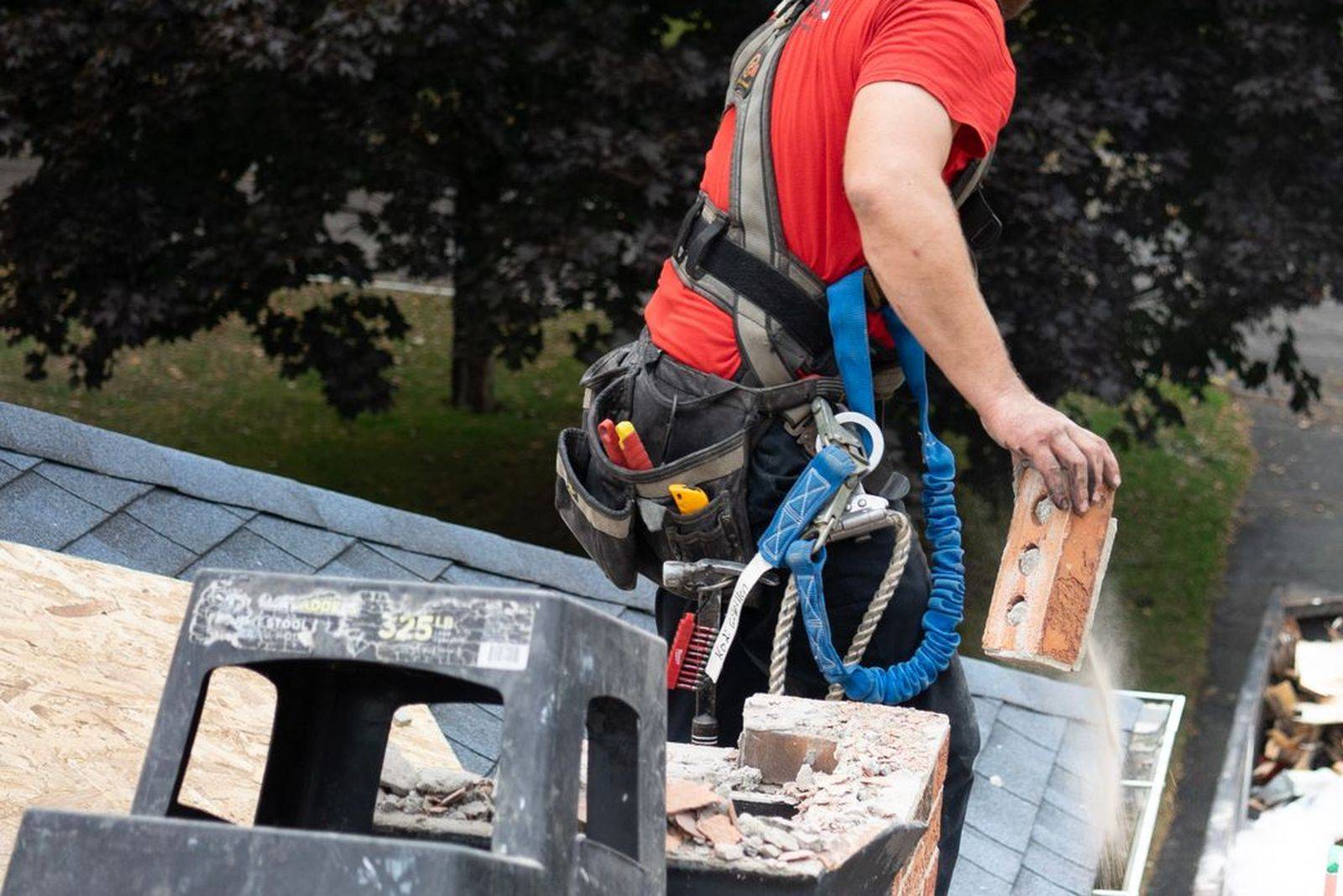Worker in a safety harness on a residential roof dropping old brick and roofing debris into a dump trailer below