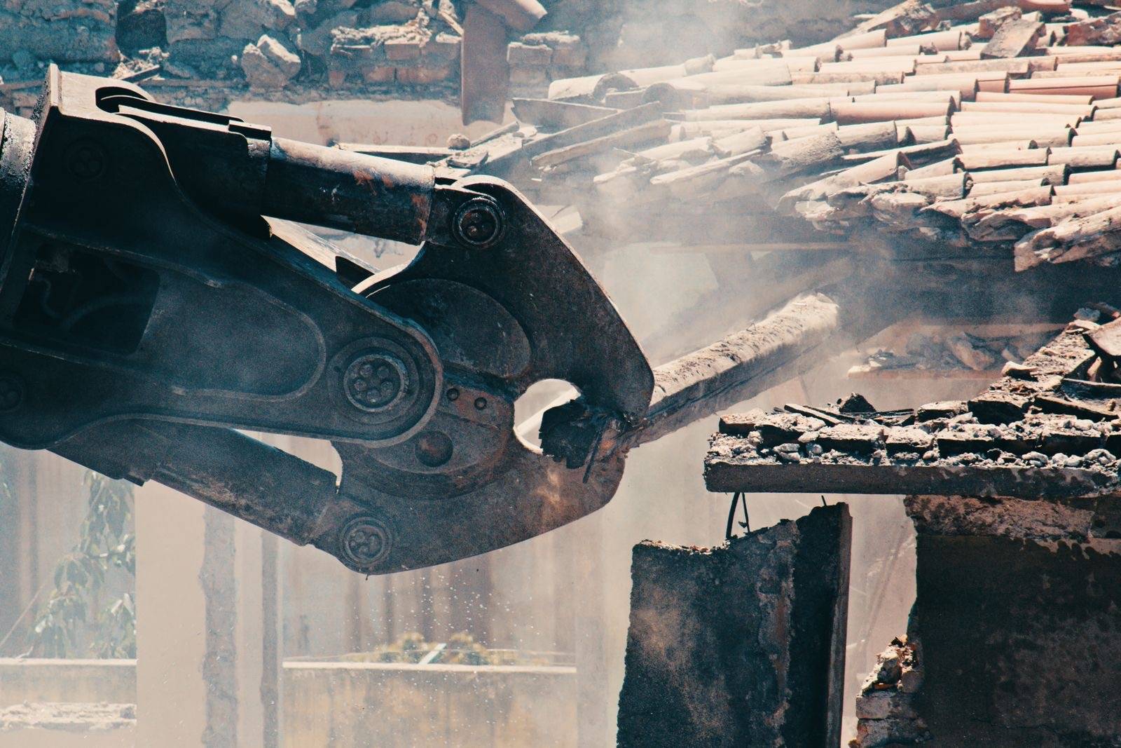 Close-up of a hydraulic demolition shear on an excavator arm biting through a roofline as dust and debris fall away
